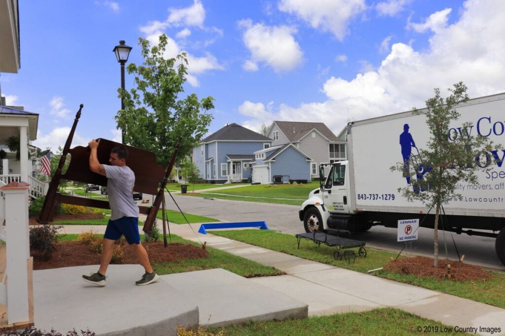 Low Country Moving Specialists crew member carrying a wooden headboard along a sidewalk past the company moving truck