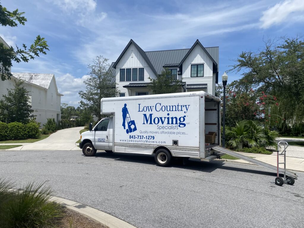 Low Country Moving Specialists box truck parked outside a white two-story house, ramp down and a hand truck beside it