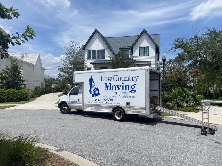 Low Country Moving Specialists box truck parked outside a white two-story house, ramp down and a hand truck beside it